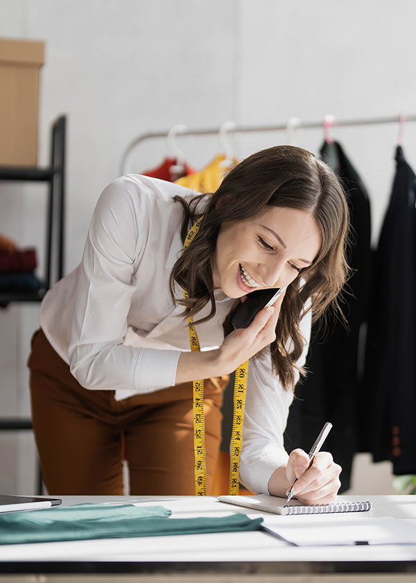 Mulher empresária ao telefone debruçada sobre a mesa para fazer anotações em um tablet. Ao fundo, roupas penduradas em uma arara.